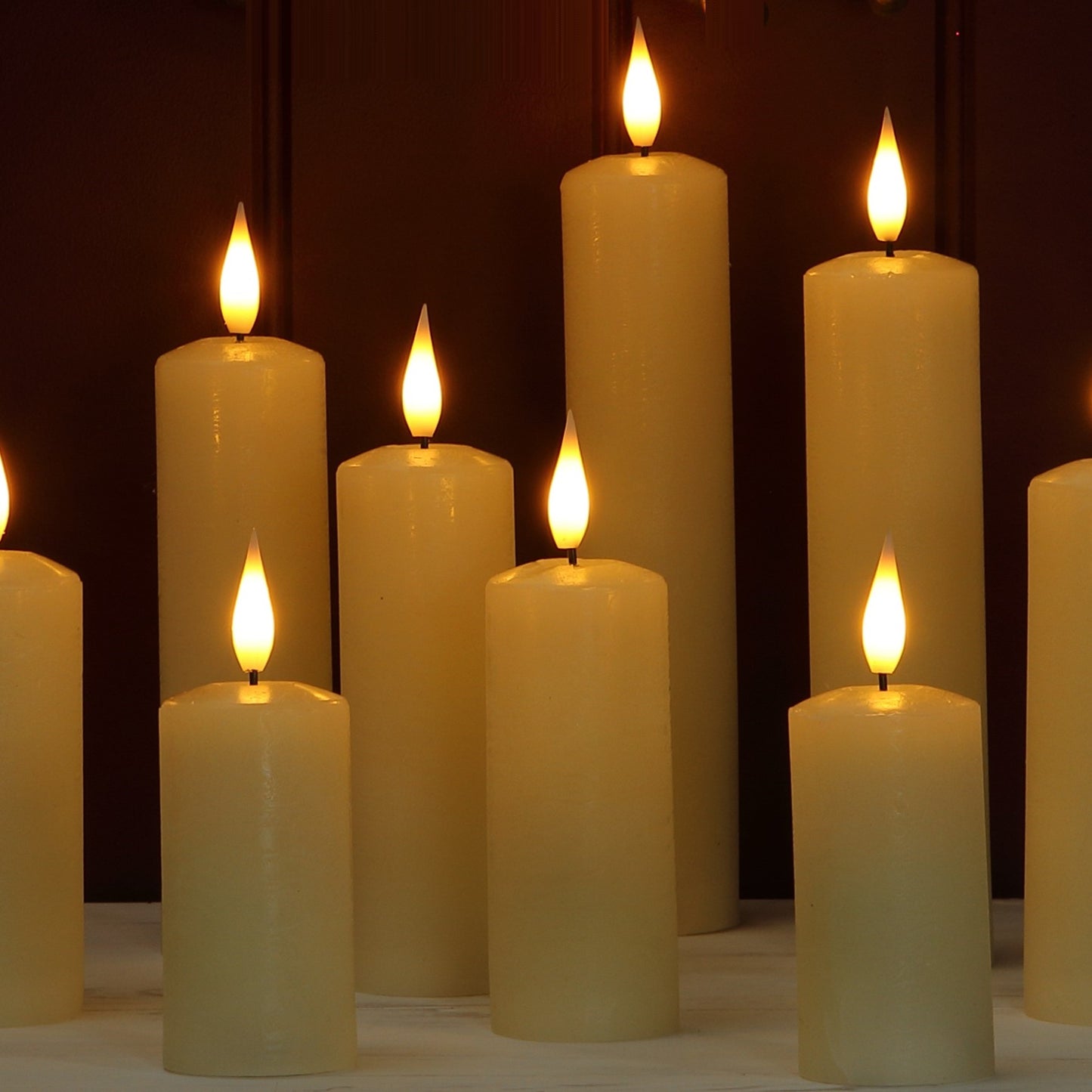 A group of cream-colored pillar candles with lit flame tops, arranged on a light surface against a dark brown background.