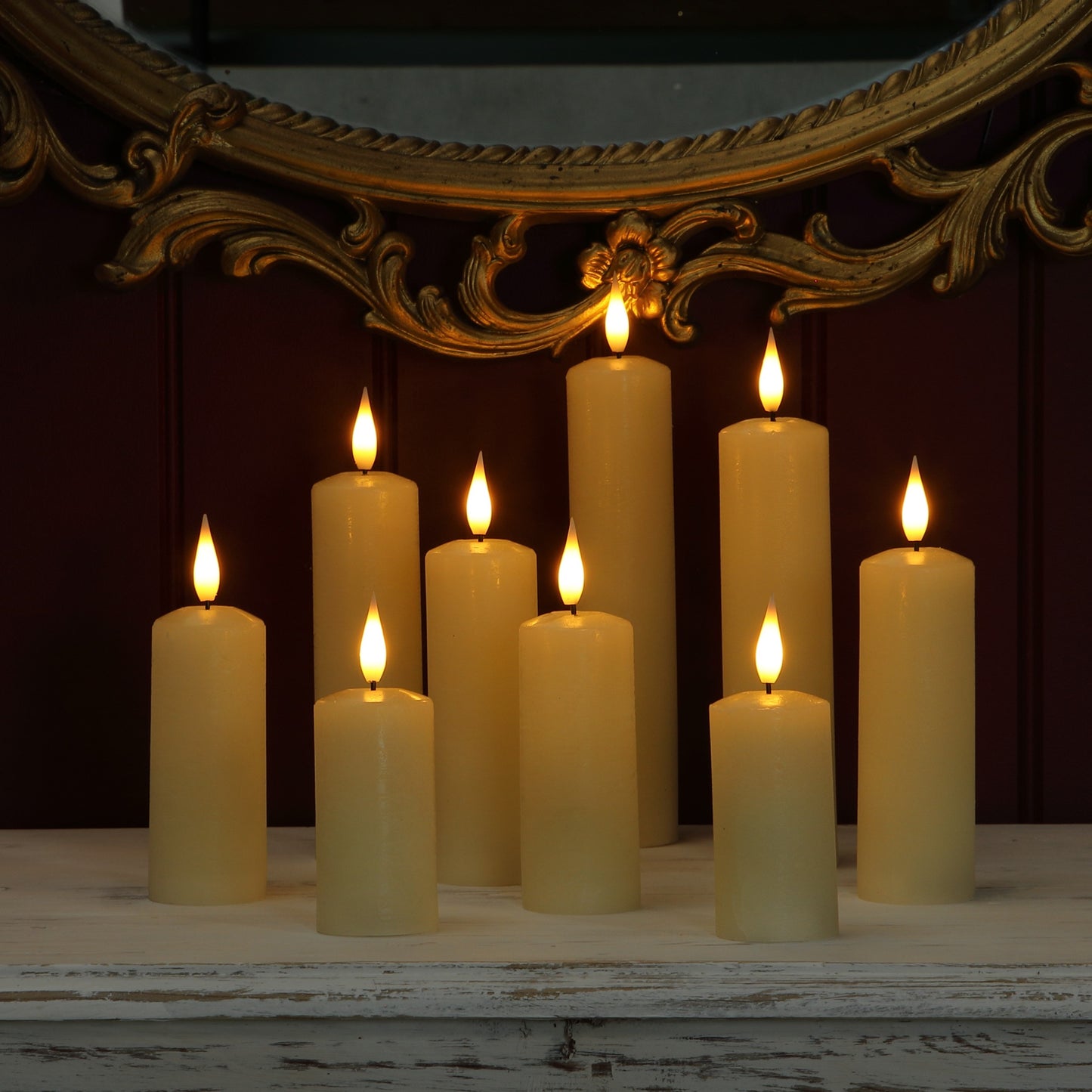 A set of nine faux pillar candles with lit flame tops arranged on a distressed white wooden mantel in front of a dark red wall and an ornate gold mirror.