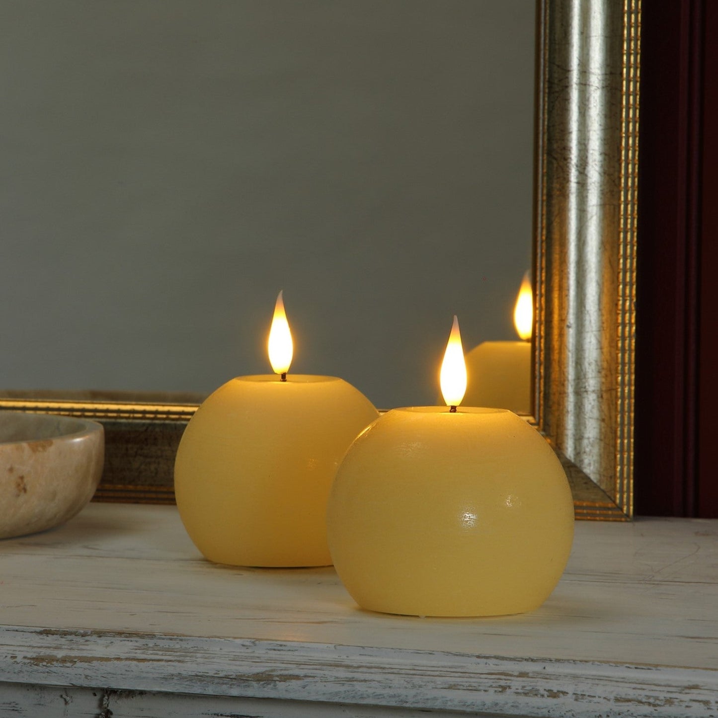Two round yellow wax candles with lit flames sit on a distressed white wooden surface in front of a mirror with a gold frame.