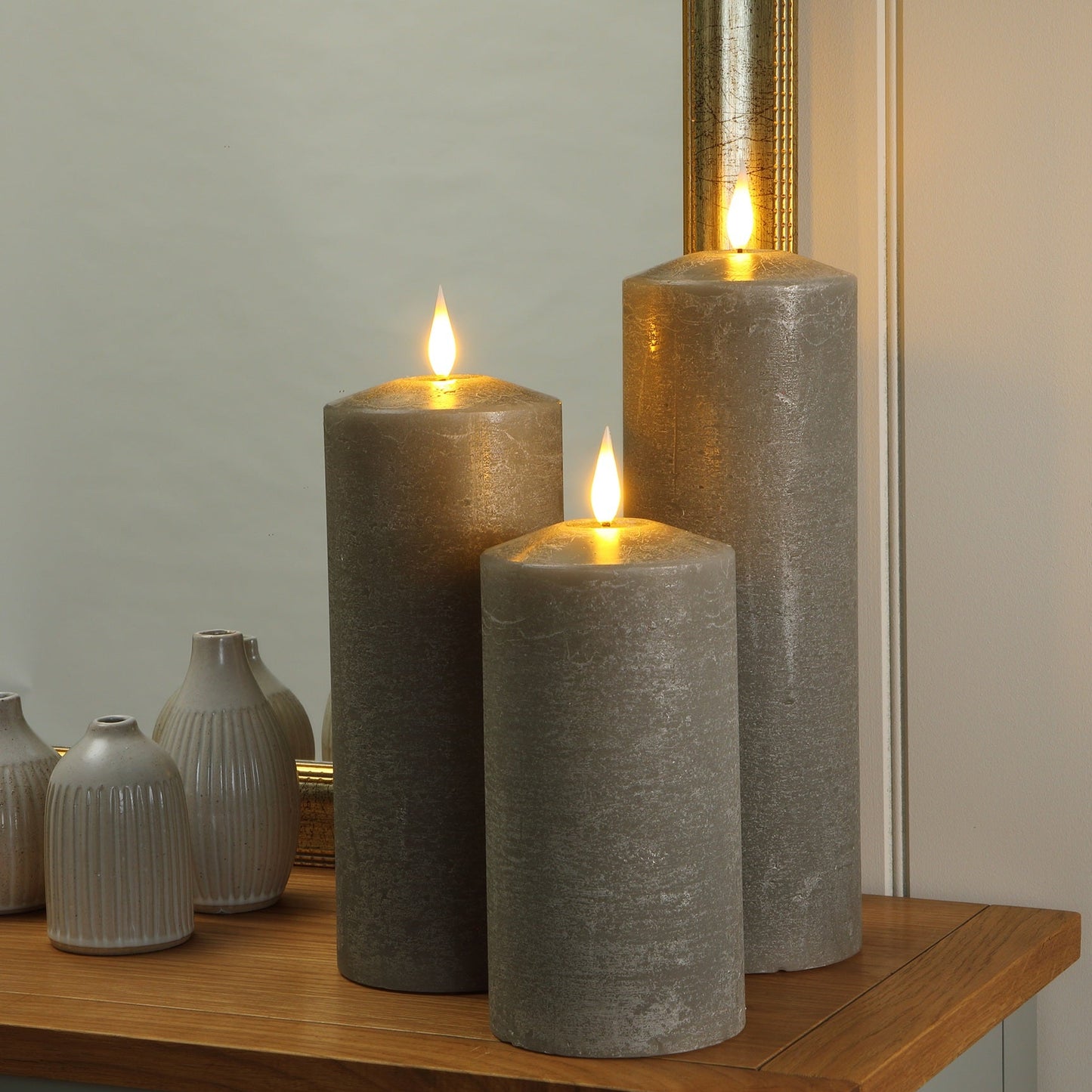 Three grey textured pillar candles with lit flames are arranged on a wooden cabinet next to small white ribbed vases in front of a mirror.