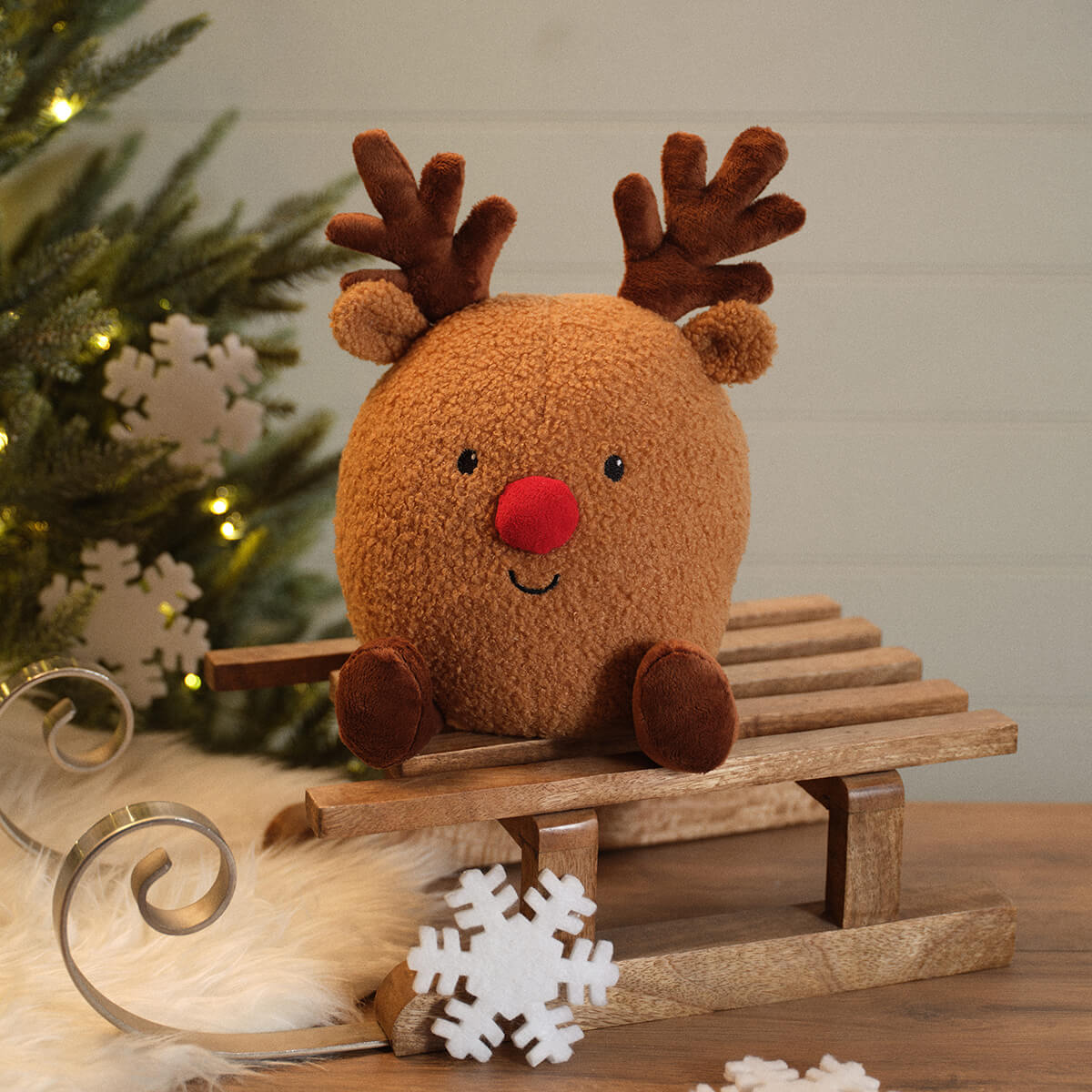 A round brown plush reindeer door stop with a red nose and dark brown antlers sits on a small wooden sled. The scene is styled with a Christmas tree, white snowflake decorations, and a warm holiday background.