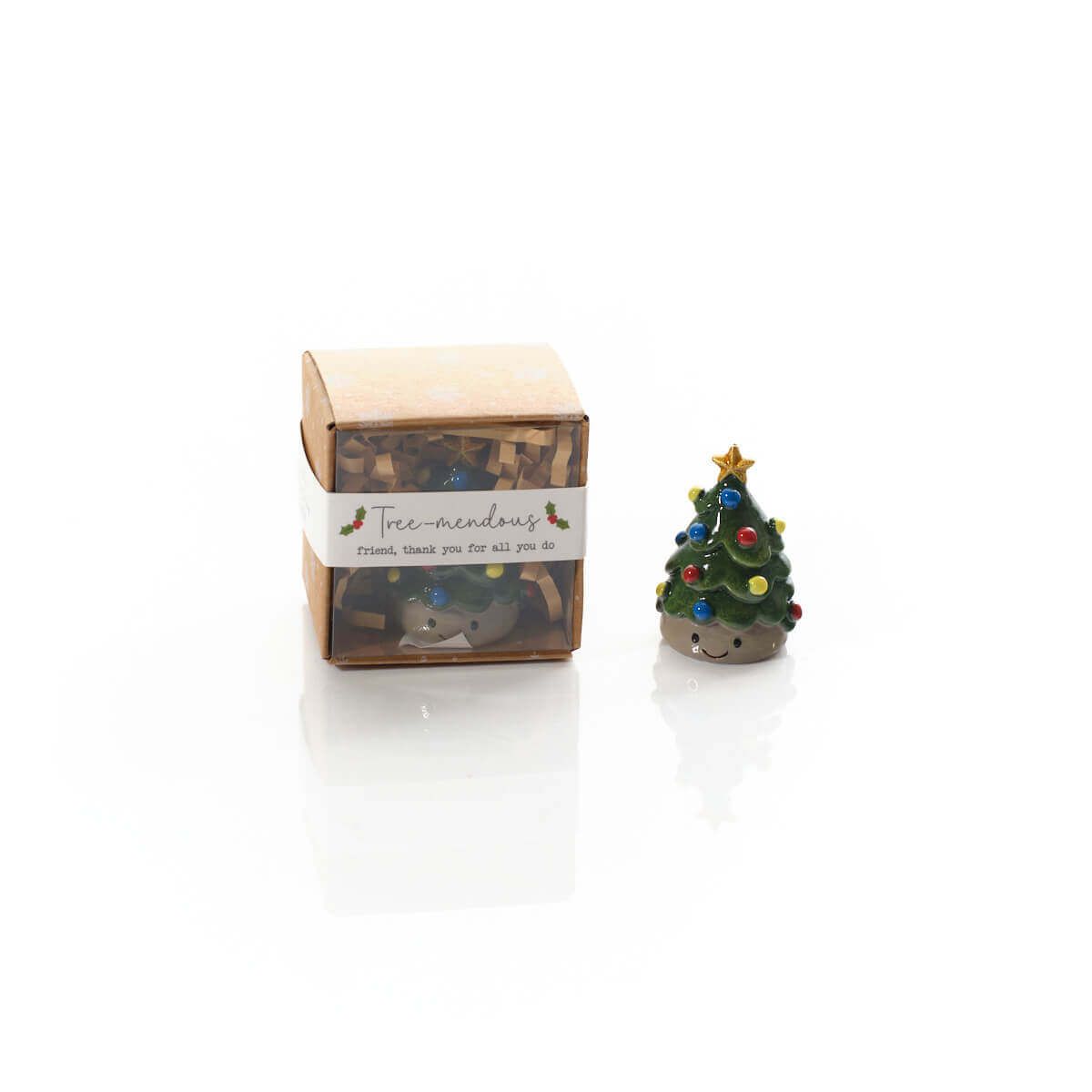 A small decorative Christmas tree figurine with colourful ornaments and a gold star stands beside its kraft paper gift box on a white reflective surface. The box has a white label reading 'tree-mendous friend, thank you for all you do'