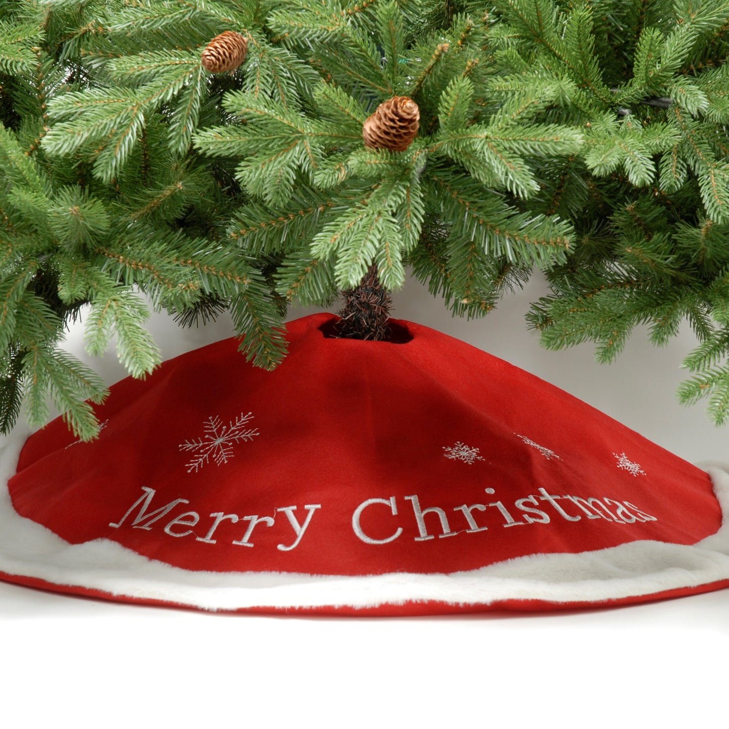 A red Christmas tree skirt with 'Merry Christmas' text and silver embroidered snowflakes, placed under a green Christmas tree with pine cones on its branches.