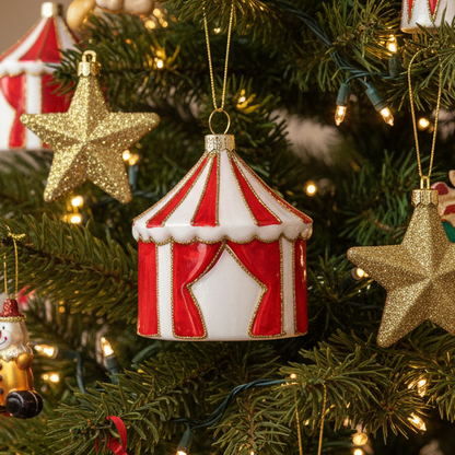 A red and white striped circus tent Christmas ornament hanging on a green Christmas tree with warm string lights and gold star ornaments.