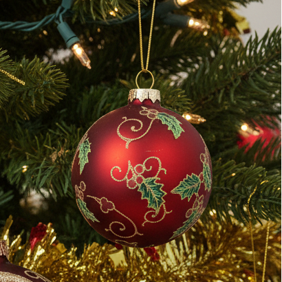 A red Christmas ornament with green holly leaves and gold swirl patterns hanging on a green Christmas tree with warm string lights.