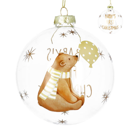 A clear round Christmas ornament featuring a watercolor-style brown bear wearing a white and yellow striped scarf, sitting beside a yellow polka-dot balloon. The ornament is decorated with gold starbursts and the words 'BABY'S FIRST CHRISTMAS' in gold.
