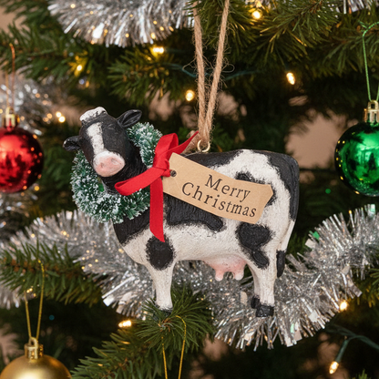A Christmas ornament shaped like a black and white cow, decorated with a green wreath and a red ribbon bow. A kraft paper tag hanging from the ribbon reads 'Merry Christmas'.