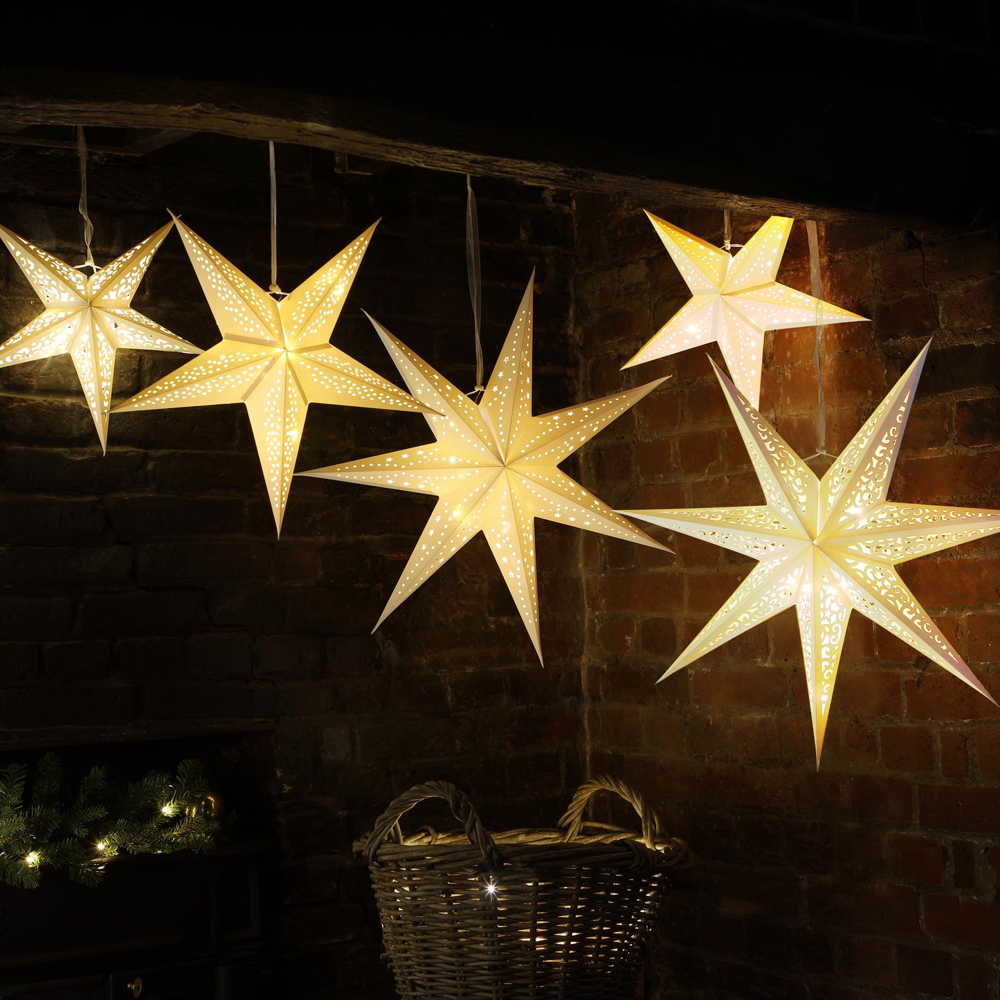 Five glowing star-shaped hanging ornaments in warm white light, displayed against a dark brick wall with holiday decor below.