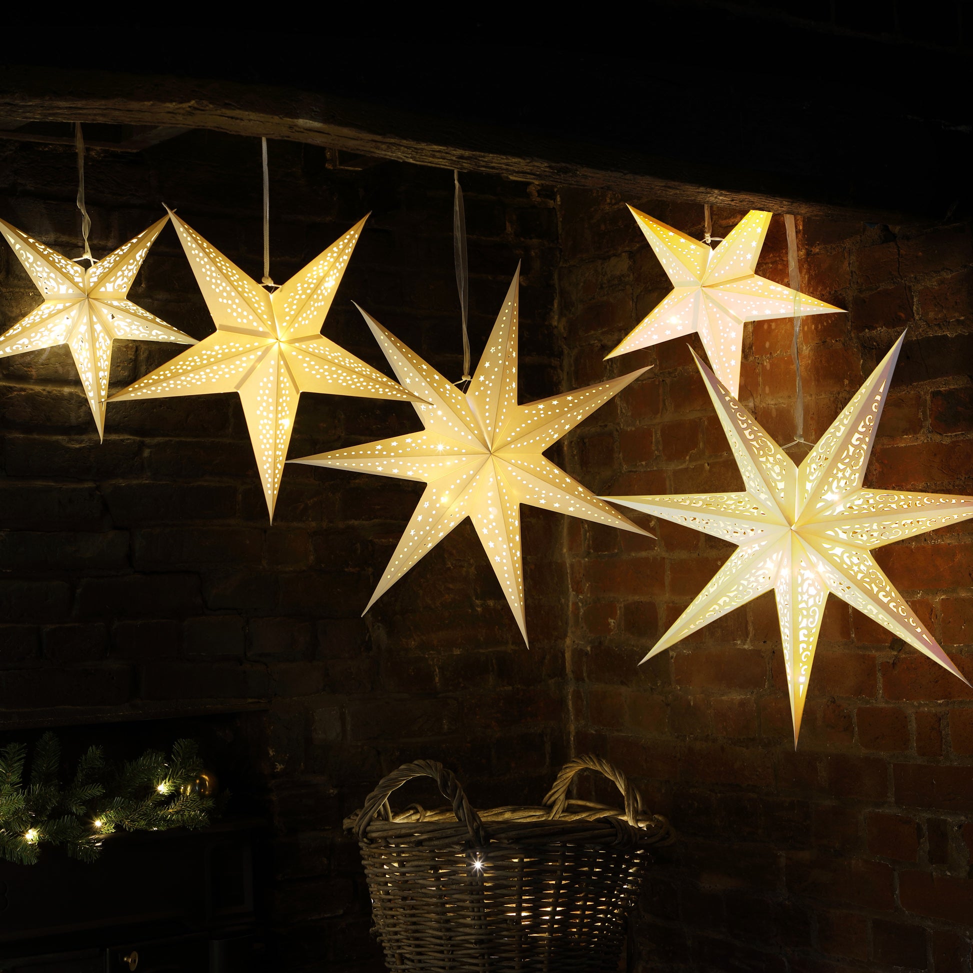 Five glowing star-shaped hanging ornaments in warm white light, displayed against a dark brick wall with holiday decor below.