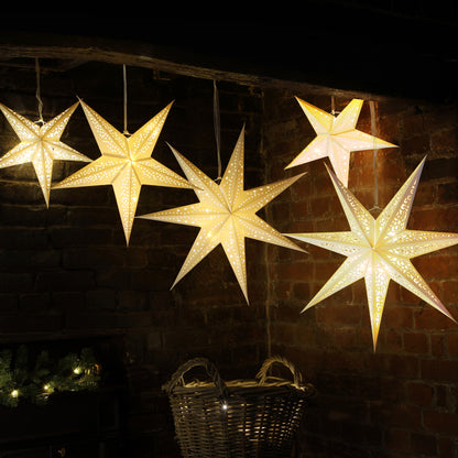 Five glowing star-shaped hanging ornaments in warm white light, displayed against a dark brick wall with holiday decor below.