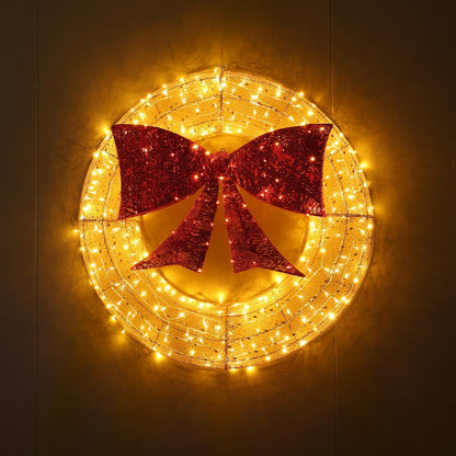 A circular holiday wreath decoration with warm white string lights and a large red glitter bow in the center, shown against a dark background.