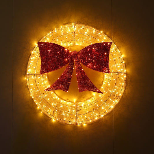 A circular holiday wreath decoration with warm white string lights and a large red glitter bow in the center, shown against a dark background.