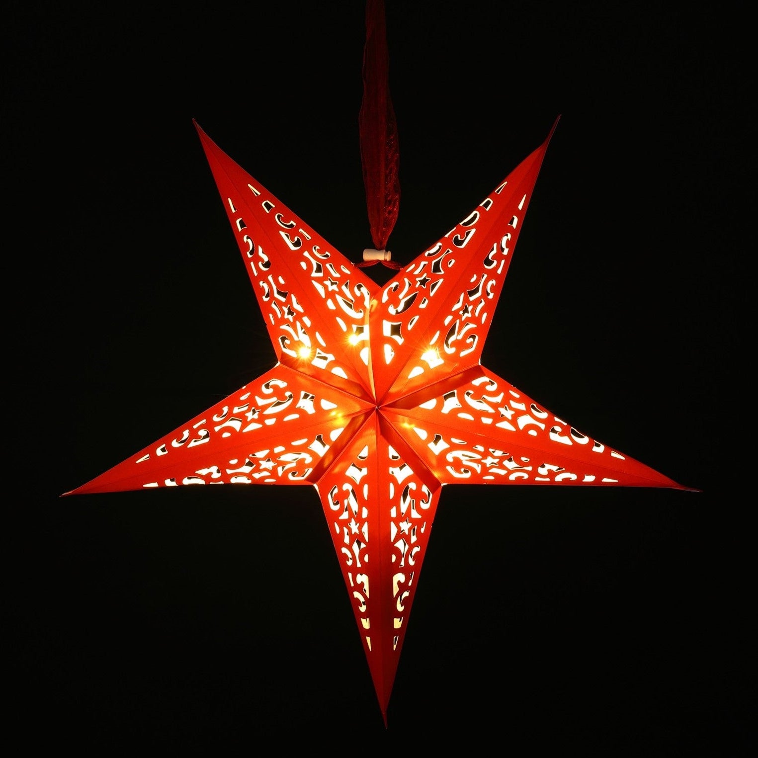 A red five-pointed star-shaped hanging ornament with intricate cutout patterns, glowing with warm light against a solid black background.