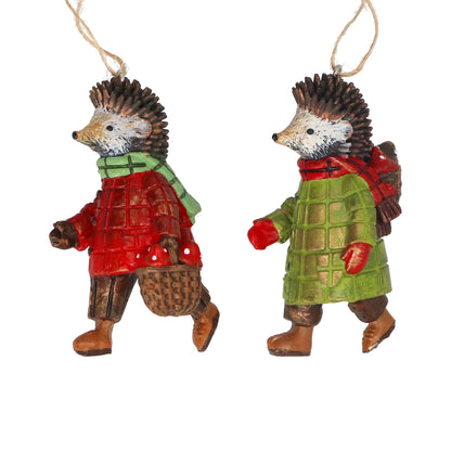 Two small hedgehog ornaments hanging from twine, dressed in winter clothing. One hedgehog wears a red coat with a green scarf and brown pants, and the other wears a green coat with red mittens and brown pants, both on a plain white background.