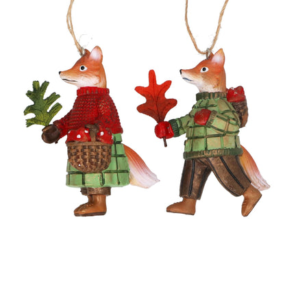 Two fox ornaments wearing sweaters, one holding a basket and the other an oak leaf, on a white background.