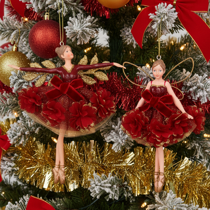 Two Christmas fairy ballerina ornaments hanging on a decorated Christmas tree. The ornaments feature miniature ballerina figures with outstretched arms, wearing red tutus with large red flowers, gold glitter accents, and gold wings, suspended by gold strings.