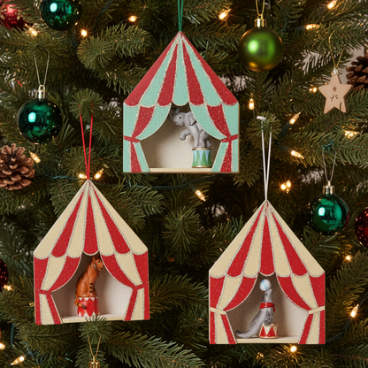 Three wooden circus tent Christmas ornaments with red and cream stripes hang on a lit Christmas tree. One tent features a gray elephant on stage, while the other two feature small figurines inside.