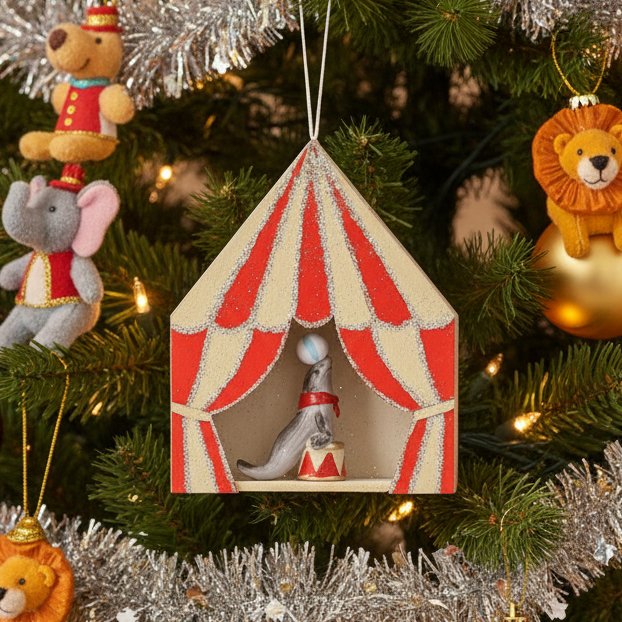 A small hanging circus-themed ornament shaped like a tent with red and cream striped curtains, glittery trim, and a figurine of a gray seal balancing a ball on its nose inside.
