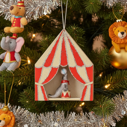 A small hanging circus-themed ornament shaped like a tent with red and cream striped curtains, glittery trim, and a figurine of a gray seal balancing a ball on its nose inside.