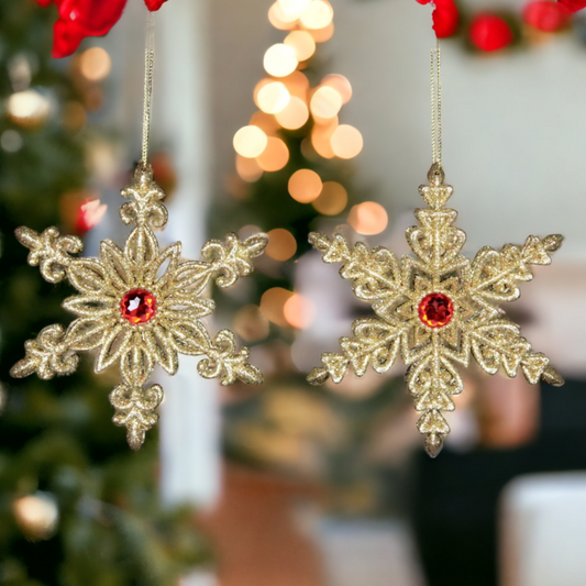Two golden glitter snowflake Christmas decorations with red jewels and gold hanging wire, displayed against a blurred Christmas tree and lights.