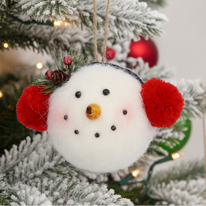 A felt snowman Christmas ornament with red pom-pom ears, a yellow nose, black eyes and mouth, and a pine branch with berries on top, hanging by a twine string against a white Christmas tree background.