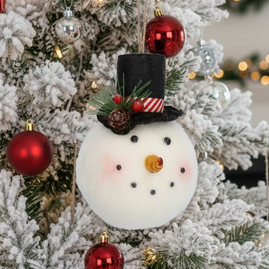 A white felt snowman Christmas ornament with a black top hat, red and white striped band, pinecones, green pine needles, and a small orange nose, hanging against a white Christmas tree background.