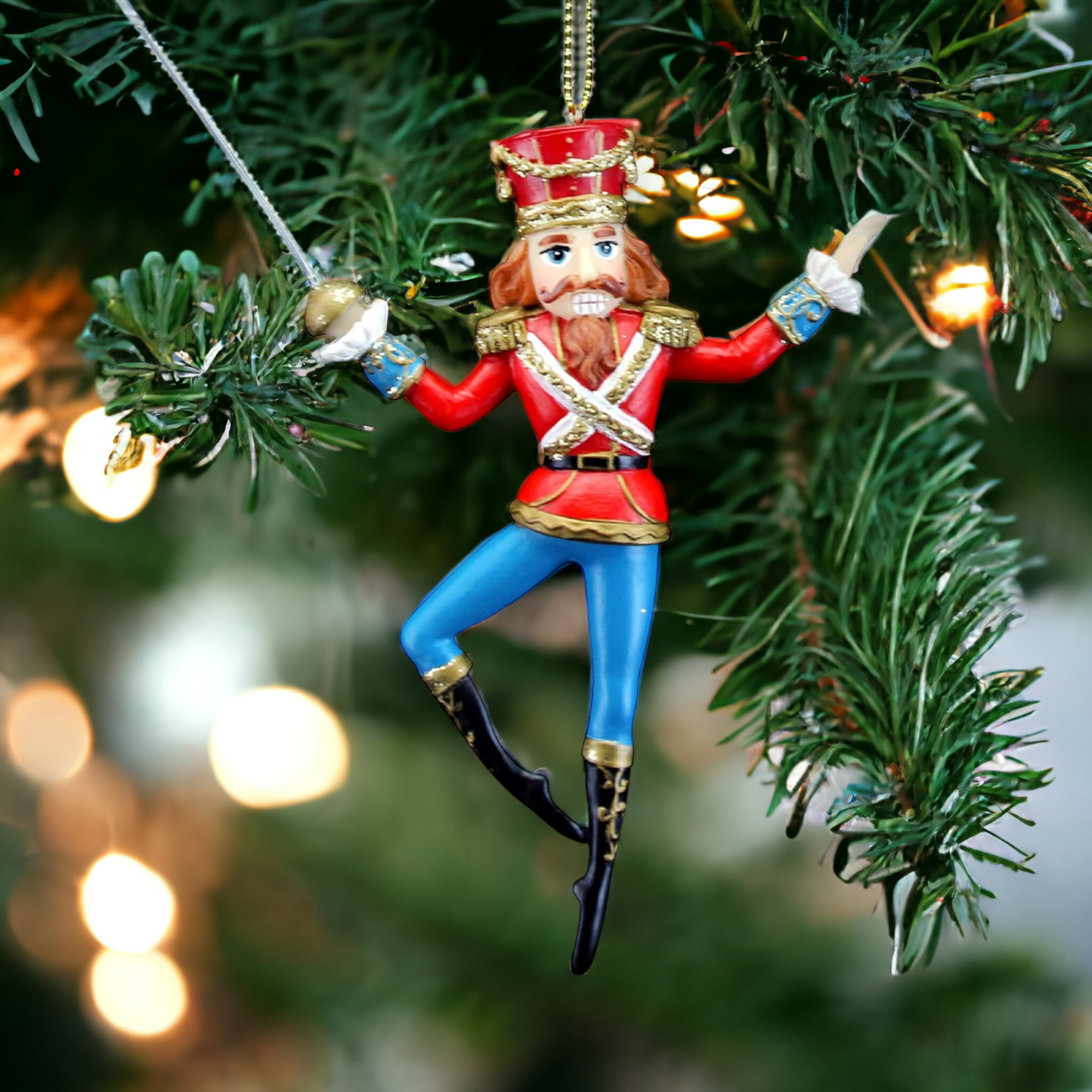 A decorative nutcracker figurine in Victorian attire, hanging from a Christmas tree. The decoration is predominantly red, with blue pants and black boots, wearing a black top hat and a sword.