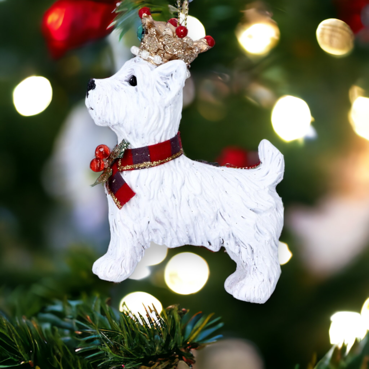 A white West Highland Terrier Christmas tree decoration with a gold crown and tartan collar, hanging from a tree with holly and Christmas lights in the background.