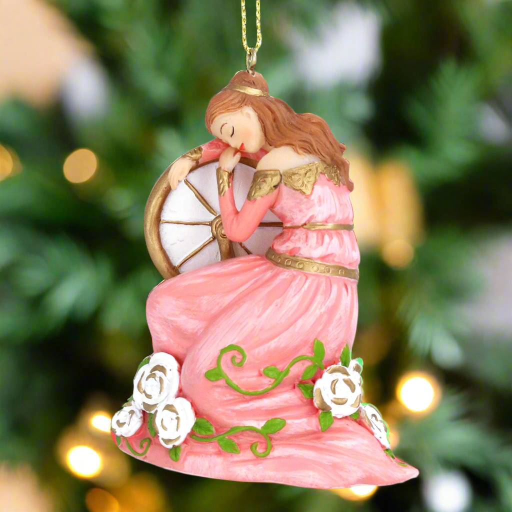 A Christmas tree decoration depicting a sleeping beauty in a pink dress, seated beside a wheel, with gold hanging wire, against a blurred background of a Christmas tree.
