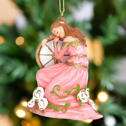 A Christmas tree decoration depicting a sleeping beauty in a pink dress, seated beside a wheel, with gold hanging wire, against a blurred background of a Christmas tree.