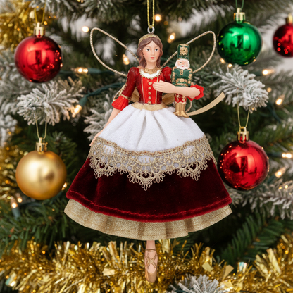 A Christmas fairy ornament hanging on a decorated Christmas tree. The ornament depicts a fairy with gold wings, a red and white dress with gold lace trim, and holds a nutcracker.