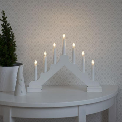 A white 7 bulb LED Christmas candle bridge battery operated, placed on a white surface with a patterned wall in the background and a small potted plant to the left.