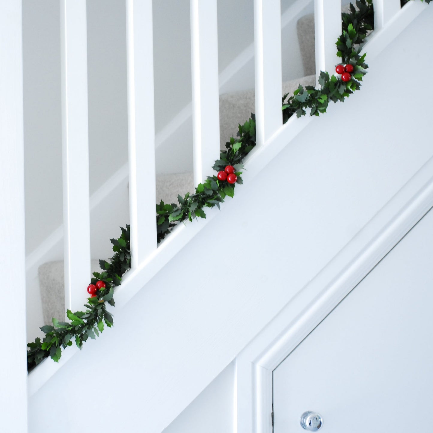 A Christmas tinsel garland with green holly and red berries decorations laid out on a white staircase, leading up to a door.