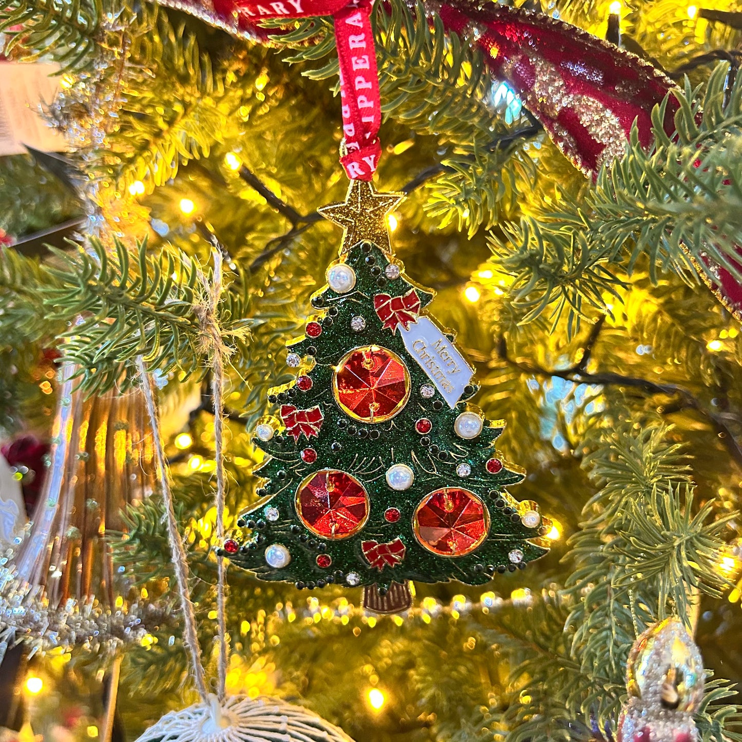 Decorative Christmas tree ornament with red and gold details hanging on a lit Christmas tree.