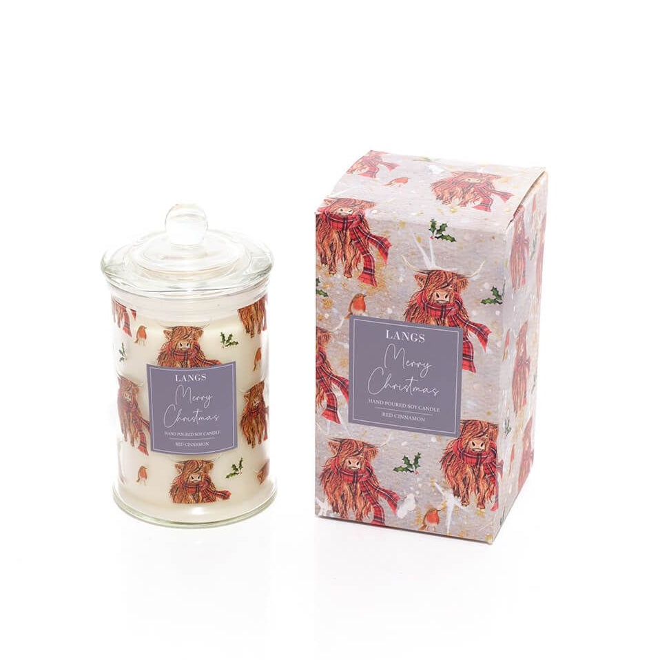 A Langs 'Merry Christmas' scented candle in a clear glass jar with a lid, shown next to its matching highland cow patterned gift box on a white background.