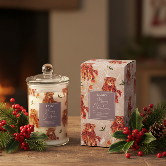 A Langs 'Merry Christmas' scented candle in a glass jar with a lid, displayed next to its patterned gift box on a wooden table decorated with pine branches, red berries, and pinecones, with a warm fireplace in the background.
