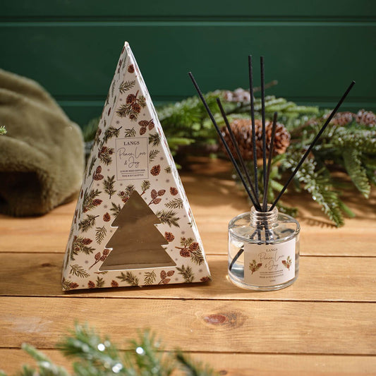 A boxed diffuser and matching reed diffuser bottle on a wooden surface, styled with evergreen branches and a green wall in the background. The triangular box and bottle label read Lana's Pine Cone & Soy.