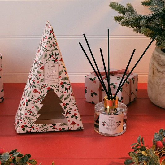 Langs Christmas Scents diffuser set featuring a triangular patterned box with a tree-shaped window, next to a glass reed diffuser bottle with black reeds and a gold collar, on a red tabletop with holiday greenery.