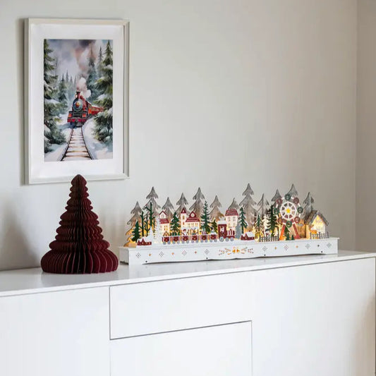 A Christmas decoration featuring a wooden structure with LED lights resembling a village, placed on a white surface, with a framed picture of a winter scene hanging above it.