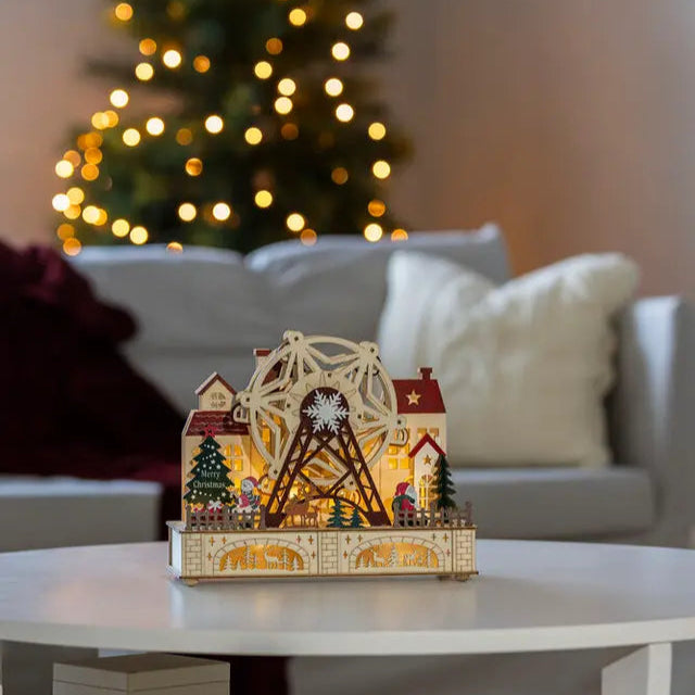 A wooden Christmas decoration featuring a rotating ferris wheel with LED lights, situated on a table with a Christmas tree and lights in the background.