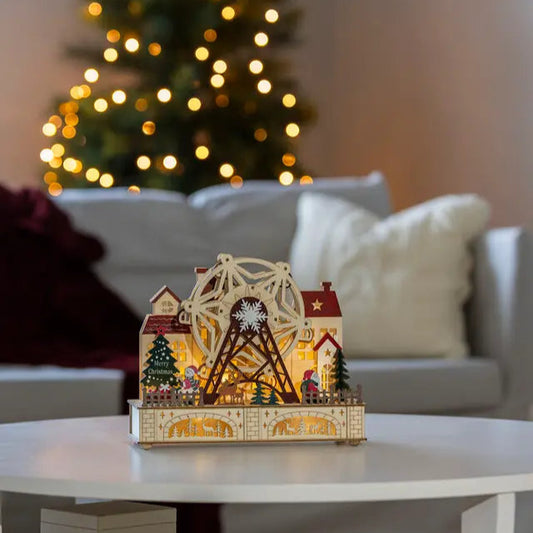 A wooden Christmas decoration featuring a rotating ferris wheel with LED lights, situated on a table with a Christmas tree and lights in the background.
