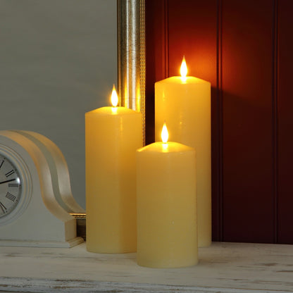 Three lit cream-colored pillar candles standing on a distressed white wooden surface, with a vintage-style clock and a dark paneled wall in the background.