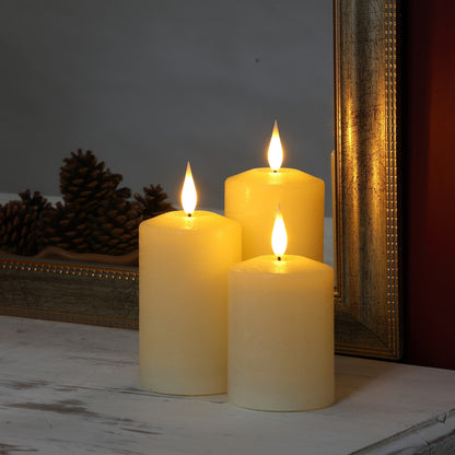 Three lit cream pillar candles arranged on a white surface in front of a gold-framed mirror, with pinecones visible in the background.