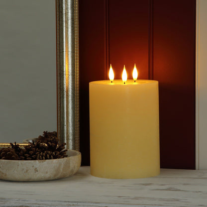 A lit three-wick pillar candle in a warm cream-yellow color sits on a distressed white wooden table, next to a decorative bowl of pinecones and a mirror, with a dark red paneled wall in the background.