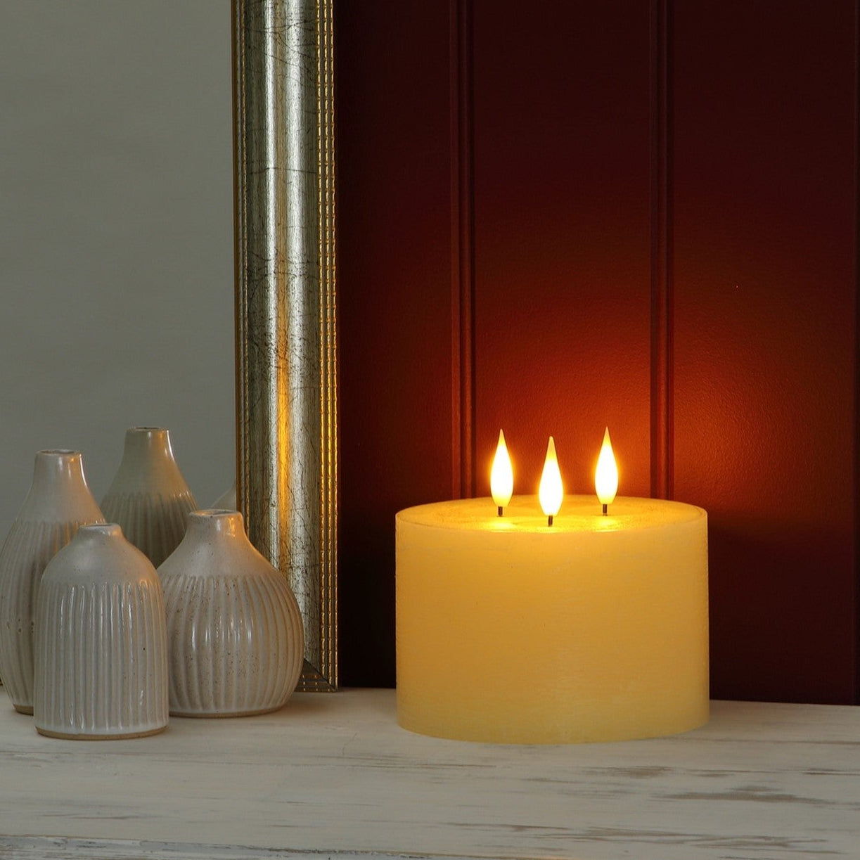 A lit three-flame pillar candle in a warm yellow glow sits on a distressed white wooden surface, with neutral-toned decorative vases and a dark paneled background.