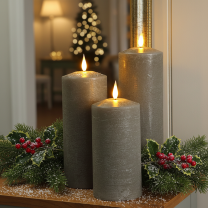 Three gray faux LED pillar candles with glowing flames are arranged on a wooden surface with green holly and pine branches decorated with red berries in a festive holiday setting.
