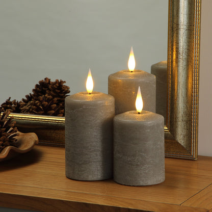 Three gray pillar candles with lit flame tops arranged on a wooden surface in front of a gold-framed mirror.