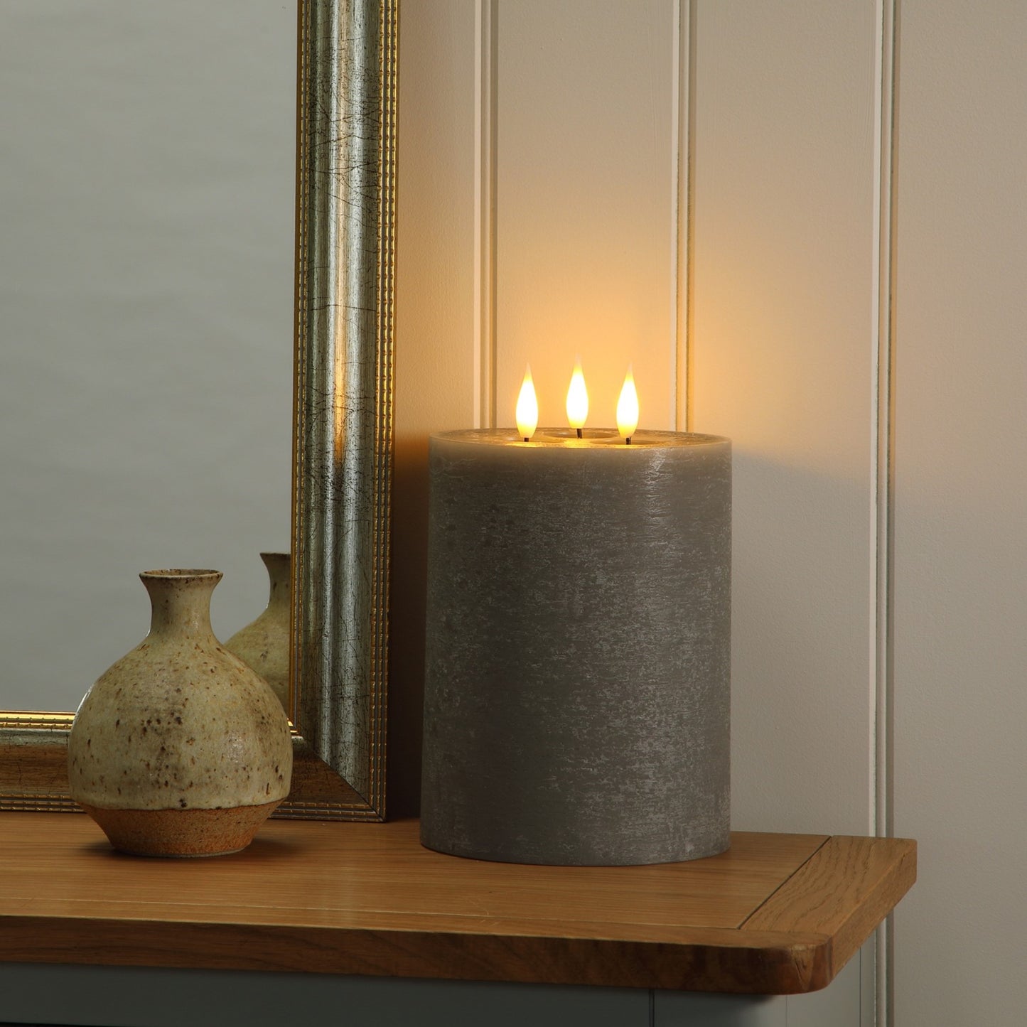 A large gray pillar candle with three lit wicks sits on a wooden shelf next to a mirror and a small ceramic vase, casting a warm glow against a paneled wall.