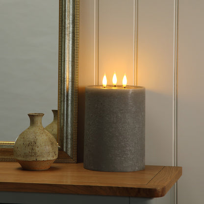 A large gray pillar candle with three lit wicks sits on a wooden shelf next to a mirror and a small ceramic vase, casting a warm glow against a paneled wall.