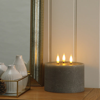 A lit grey pillar candle with three flames sits on a wooden console table next to a mirror and white decorative vases.